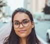 A young woman with long dark hair and glasses smiling softly while standing on a city street. The background is slightly blurred, showing cars and buildings on either side.